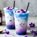 Iced butterfly pea flower tea latte in clear plastic cups with layered purple and blue hues, topped with edible flowers and black straws.