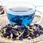 How to Make Butterfly Pea Flower Tea 5 Glass cup filled with vibrant blue butterfly pea flower tea, surrounded by dried butterfly pea blossoms on a woven mat.