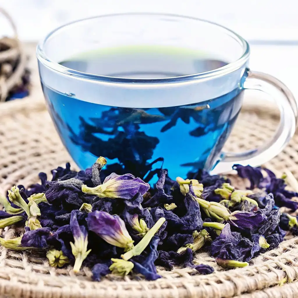 Glass cup filled with vibrant blue butterfly pea flower tea, surrounded by dried butterfly pea blossoms on a woven mat.