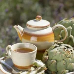 A ceramic teapot and cup filled with Artichoke Tea Vietnam, placed beside fresh artichokes in a rustic wooden box outdoors.