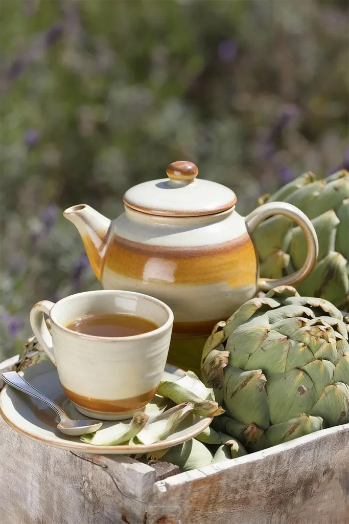 A ceramic teapot and cup filled with Artichoke Tea Vietnam, placed beside fresh artichokes in a rustic wooden box outdoors.