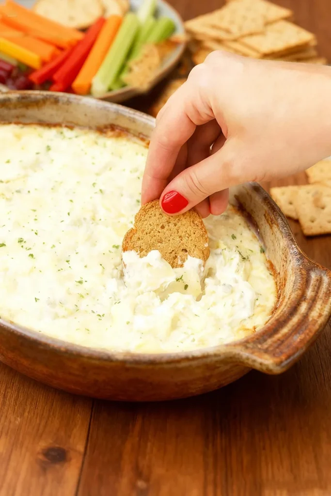 Gluten-Free Artichoke Dip being scooped with a gluten-free cracker, surrounded by colorful vegetable sticks and crackers.