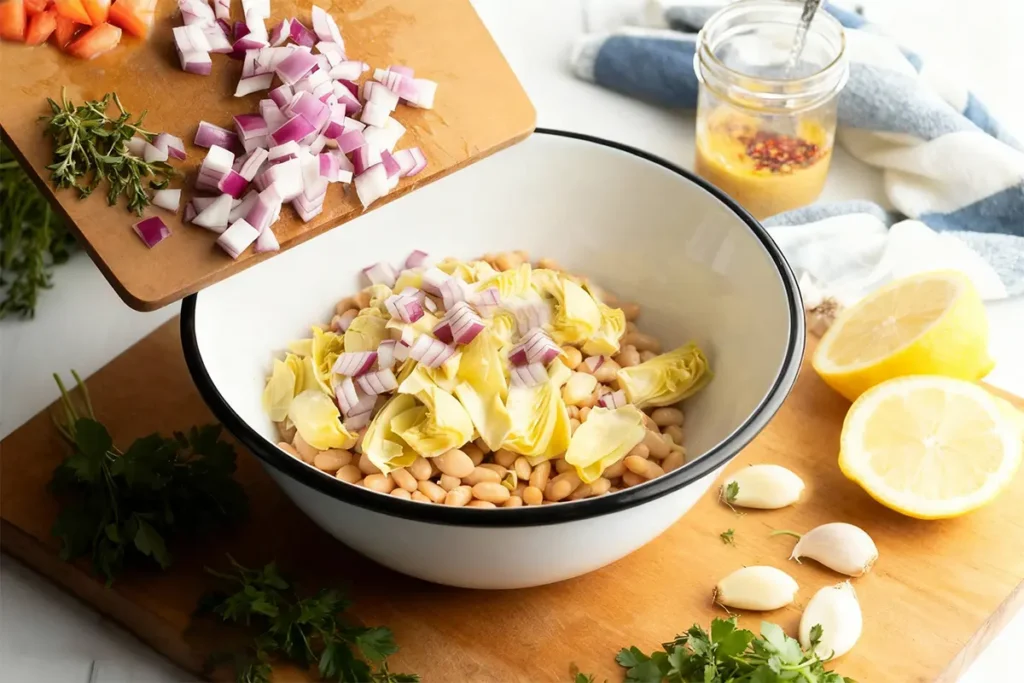 Mediterranean Artichoke and Bean Salad 6 Artichoke and bean salad ingredients being prepared: chopped red onions added to a bowl with white beans and artichoke hearts on a wooden board with lemons, garlic, and herbs.