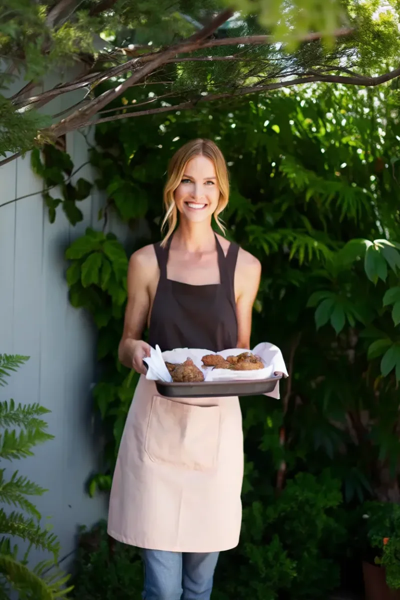 A smiling woman in a dark top and apron holding a baking tray with freshly baked goods outdoors.