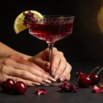 Jillian Michaels gelatin anti-hype recipe: elegant manicured hands holding crystal coupe glass filled with deep ruby hibiscus gelatin drink garnished with lemon slice and dried hibiscus petals, fresh tart cherries and hibiscus flowers scattered on dark surface with golden bokeh