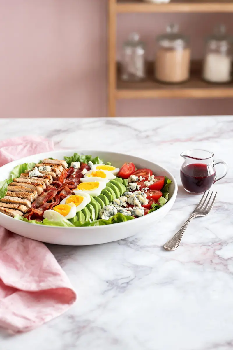 Low-carb Cobb salad in a white bowl with sliced chicken, bacon, eggs, avocado, and red wine vinaigrette on a marble table