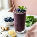 Flat belly blueberry smoothie in a tall glass topped with blueberries and chia seeds, surrounded by ginger, banana, yogurt, and spinach.